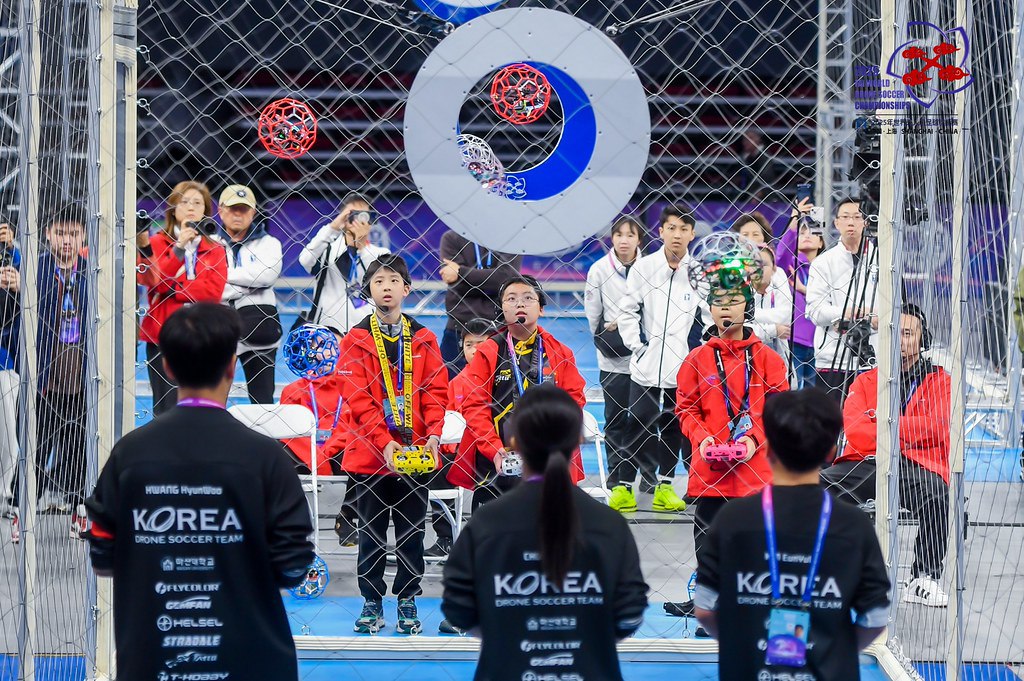 В Шанхае прошел Первый чемпионат мира по дрон футболу   В 2025 году дрон футбол включили в список спортивных дисциплин ФАИ Этот вид спорта объединяет высокие технологии и физическую активность требуя от участников технических навыков и моторики Механика игры Матчи проводятся в три сета каждый из которых длится три минуты Команда забившая больше голов в сете выигрывает этот сет Команда первой выигравшая два сета выигрывает матч При результате ничья назначается серия пенальти У каждой команды есть три попытки а у нападающего есть 10 секунд чтобы пробить пенальти в ворота Защитник команды соперника пытается заблокировать бросок Если после серии пенальти счёт остаётся равным проводится повторная серия пенальти Первое и второе место заняли команды из Китая а третье участники из Южной Кореи На соревнованиях было представлено 46 команд из 18 стран в числе которых были и Япония Франция Германия Сингапур Турция Австралия США Также были представлены команды из Гонконга Макао Тайваня Новая спортивная дисциплина подтверждает тренд на подготовку специалистов для авиации через инженерные соревнования В России дрон футбола пока нет хотя проводятся многие другие соревнования гонки дронов соревнования по программированию БАС и по киберавтономности мастер классы по использованию беспилотников в логистике и различные хакатоны по сценариям применения БАС Сейчас ассоциация Аэронекст выдвигает инициативу о запуске в России нового формата многодисциплинарных инженерных соревнований умных дронов DRONOSAPIENS GAMES Представители заявляют что для таких соревнований акцент может быть поставлен не столько на превосходство моторики оператора а на интеллектуальные способности участников В финальный день соревнований над рекой города провели уже традиционное под любые мероприятия для китайцев шоу дронов   Дронофлот